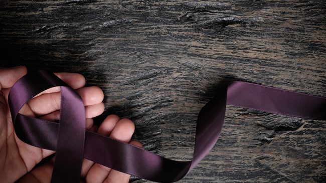 Top view of male hand holding purple ribbon on wood background with copy space. Pancreatic cancer, epilepsy, leiomyosarcoma, lupus, Alzheimer’s disease and cystic fibrosis awareness concept. Sanará Mañana - Enfermeria y Malos Tratos