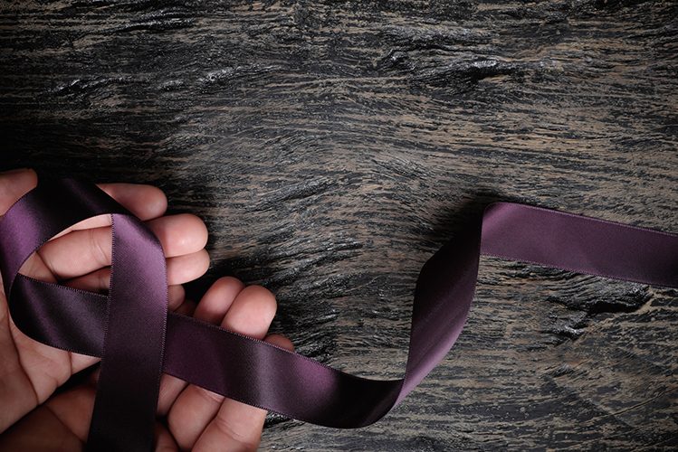 Top view of male hand holding purple ribbon on wood background with copy space. Pancreatic cancer, epilepsy, leiomyosarcoma, lupus, Alzheimer’s disease and cystic fibrosis awareness concept. Sanará Mañana - Enfermeria y Malos Tratos